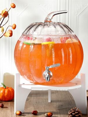 Pumpkin-shaped glass beverage dispenser with white wooden base and stainless steel spigot for parties.